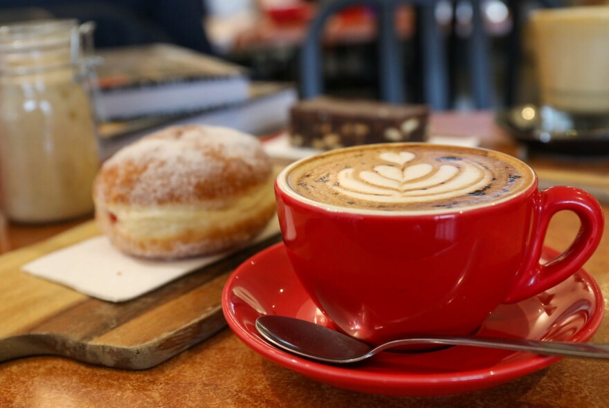 Swirled caffe latte in a red cup and saucer with a donut on a wooden board.