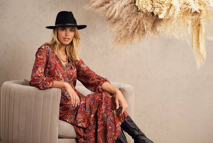 A model sitting in on a couch wearing a red paisley maxi dress, knee-high black boots and a black fedora 