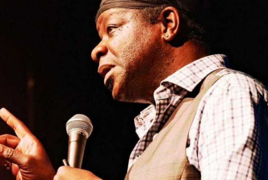 Stephen K Amos on stage, holding a microphone and looking serious.