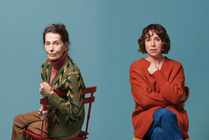 Two women, one older, one middle-aged, seated on simple chairs, looking at camera, against blue-grey background.