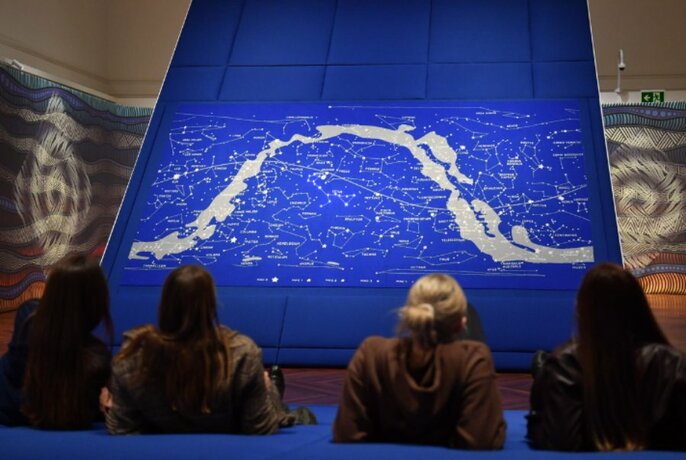 Four people reclined on soft seats looking at a large blue star map on display inside a room at the State Library.