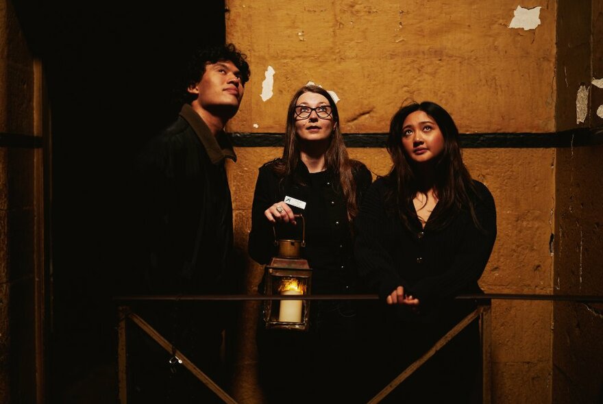 Three people inside a dimly-lit room, one holding a lantern, all looking upwards. 