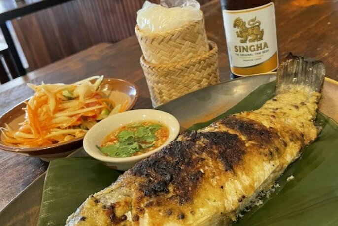 Whole grilled fish served on a large banana leaf, with a bowl of salad and a bottle of beer in the background.