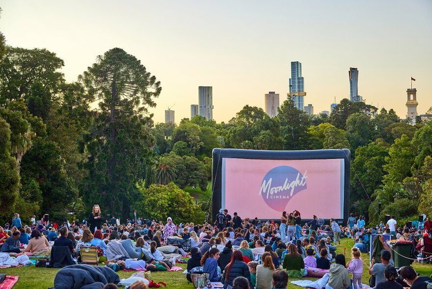 A giant movie screen in a city park with a large audience sitting on the grass.