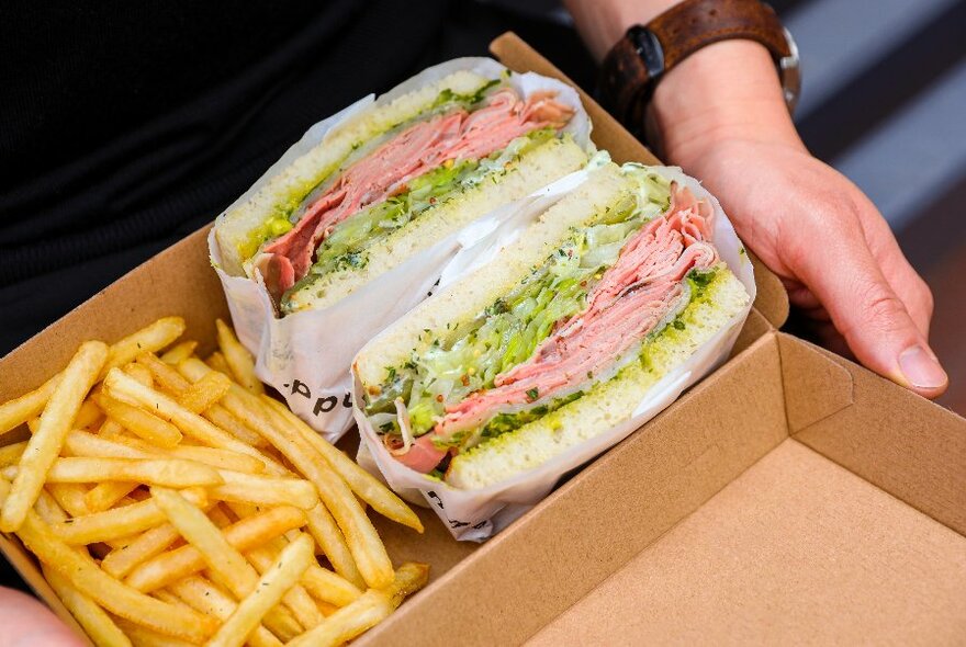 Generously-filled sandwich next to chips in a cardboard container, hand and wrist with watch visible. 