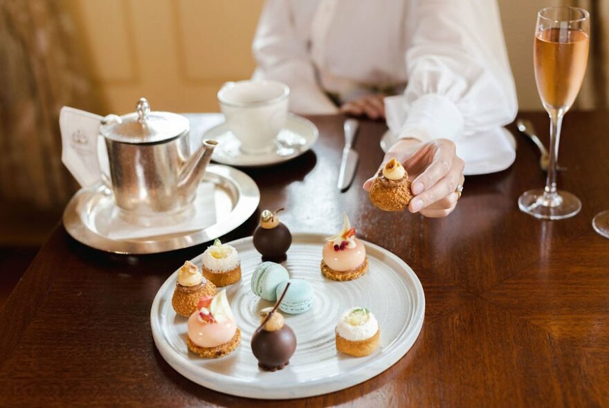 A hand reaching to select a small dessert from a plate of afternoon tea sweet treats, with a silver pot of tea, a china cup and saucer, and a glass of pink sparkling wine resting on the table.