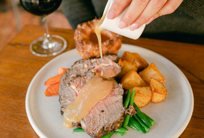 Someone pouring gravy over a roast beef dish with carrots, potatoes, beans and a Yorkshire pudding.
