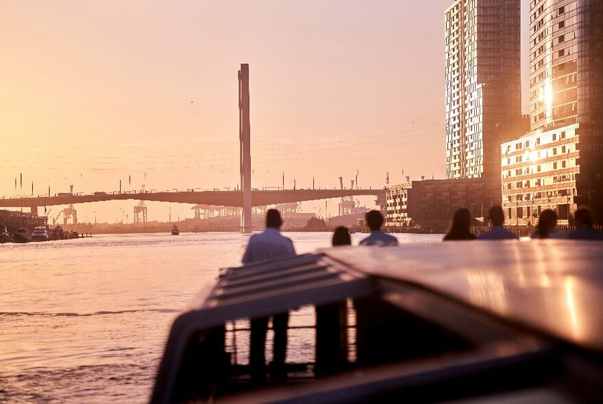 People standing at the front of a riverboat at dusk, with a bridge on the horizon and city buildings to the side.