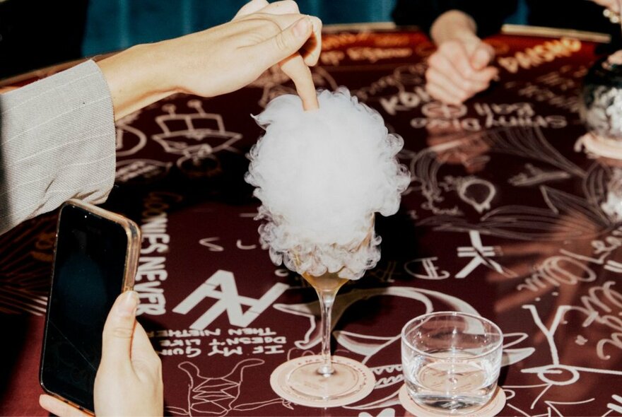 Person taking a photo on their smartphone of them putting their finger into a cocktail that is garnished with fairy floss.