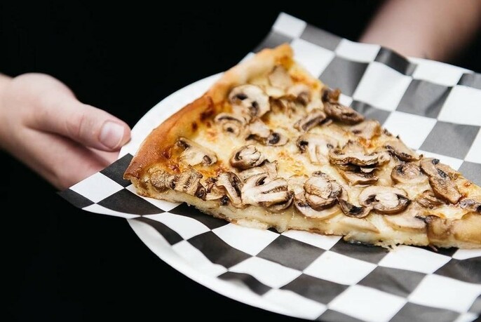 A hand holding a slice of funghi pizza on a black and white checked serviette.