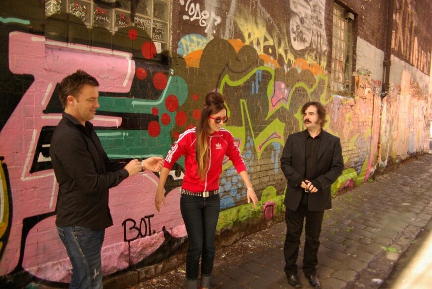 Two men and a woman posing in an outdoor laneway in front of a wall covered in street art.