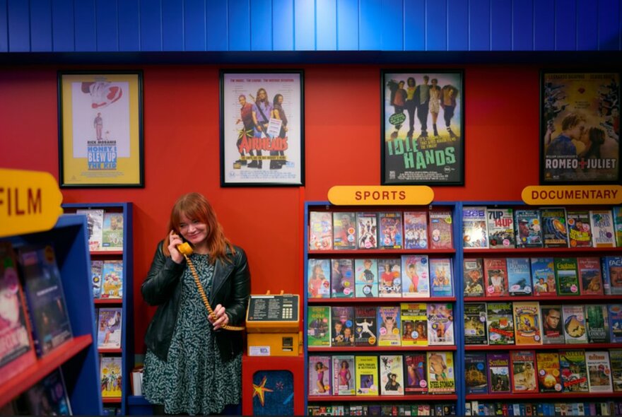 A woman on a vintage yellow pay phone amongst a colourful artistic installation of rental video tapes.