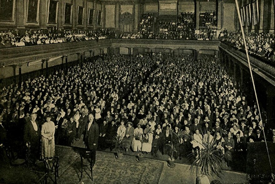 Vintage black and white photograph dating from the late 1800s, of a large assembly hall filled with a seated audience and people also seated in the balcony circle, with three people standing on the stage. 