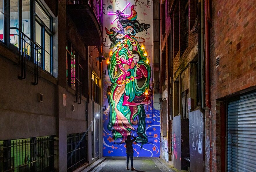 A man taking a photo of a giant street art mural in a narrow laneway at night.