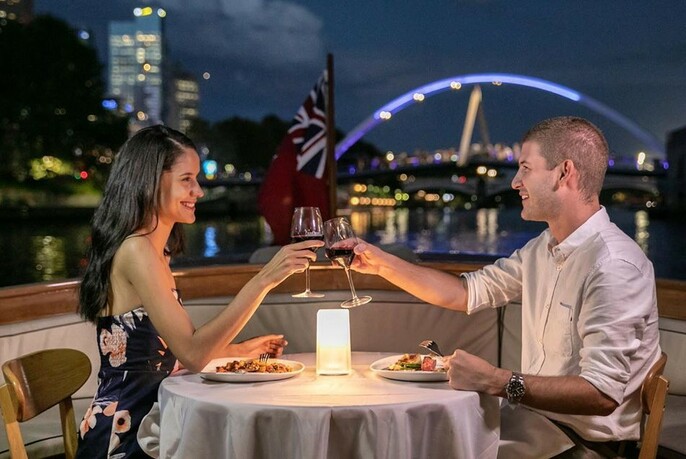 Couple eating a meal at a candlelit table on a boat with river and buildings in the background.