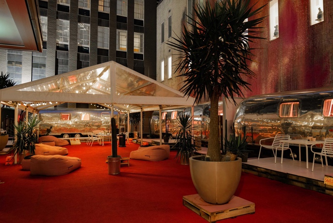 Three vintage 1970s silver airstream trailers on a red AstroTurf city building rooftop, with potted palms, bean bags and marquees.