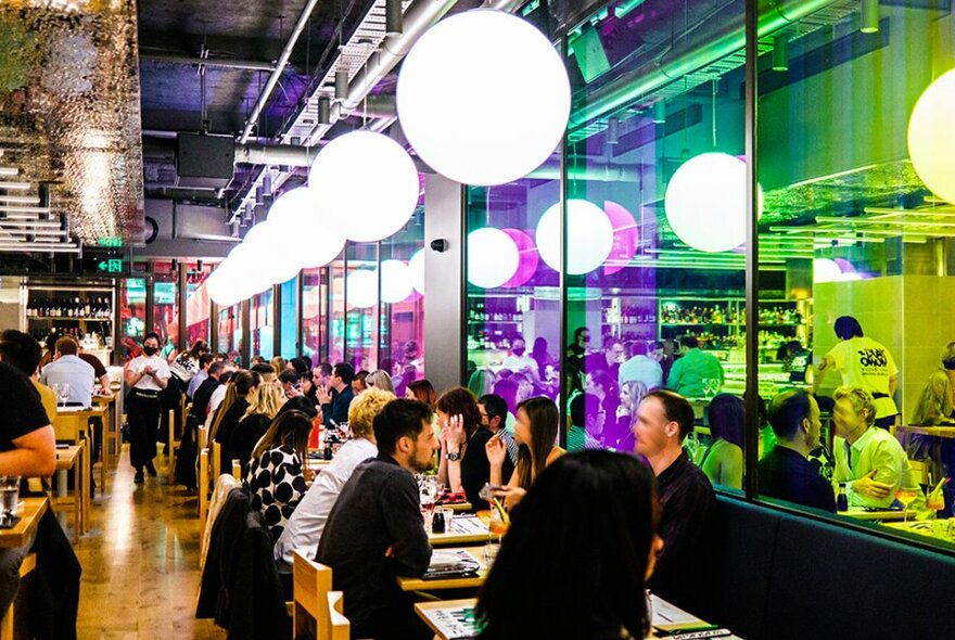 Diners in a restaurant with neon windows and large pendant lights.