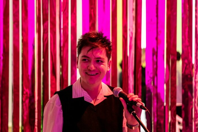 A person with short hair, talking into a microphone on a stand, in front of a pink striped background.