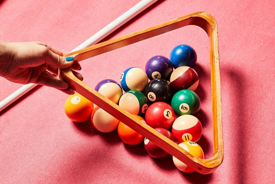 A person lifting a frame from a pink pool table with balls arranged for a game.