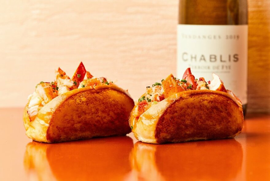 Two lobster rolls on an orange bench next to a bottle of Chablis.