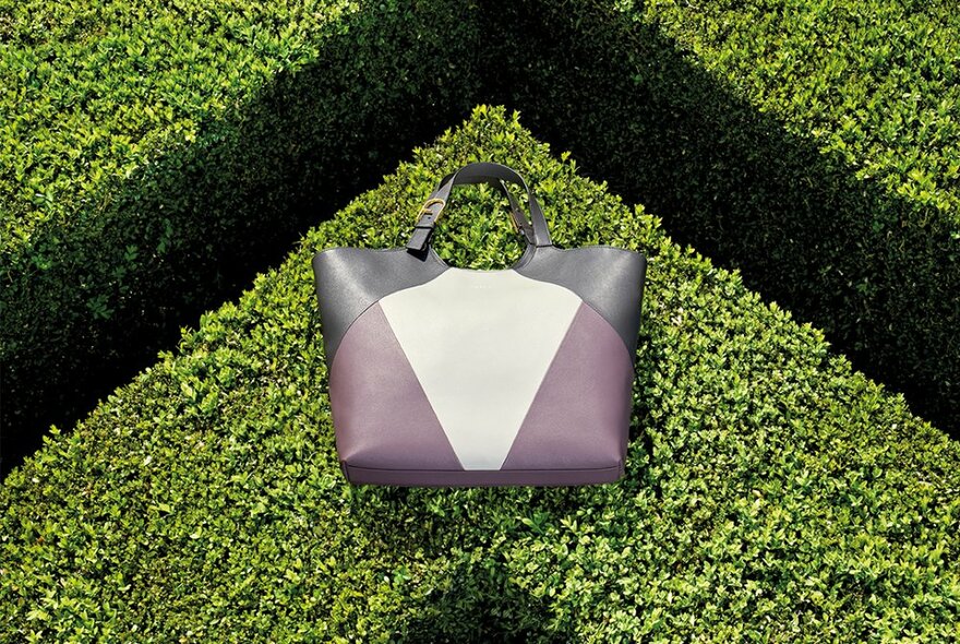 A leather tote bag made up of geometric panels of different coloured leather, resting on the top of a very neatly pruned green hedge. 
