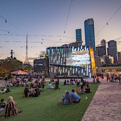 People sitting on the ground watching the Fed Square big screen in the city.