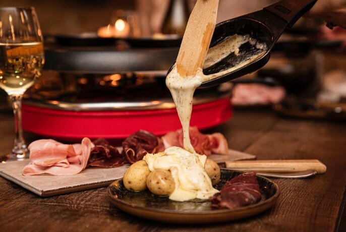 Melted cheese being poured over a plate of mixed charcuterie and boiled potatoes, a glass of white wine in the background.