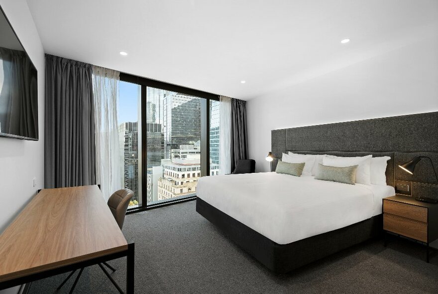 Simply furnished hotel room with grey carpet, floor-to-ceiling windows, white double bed and wooden table.