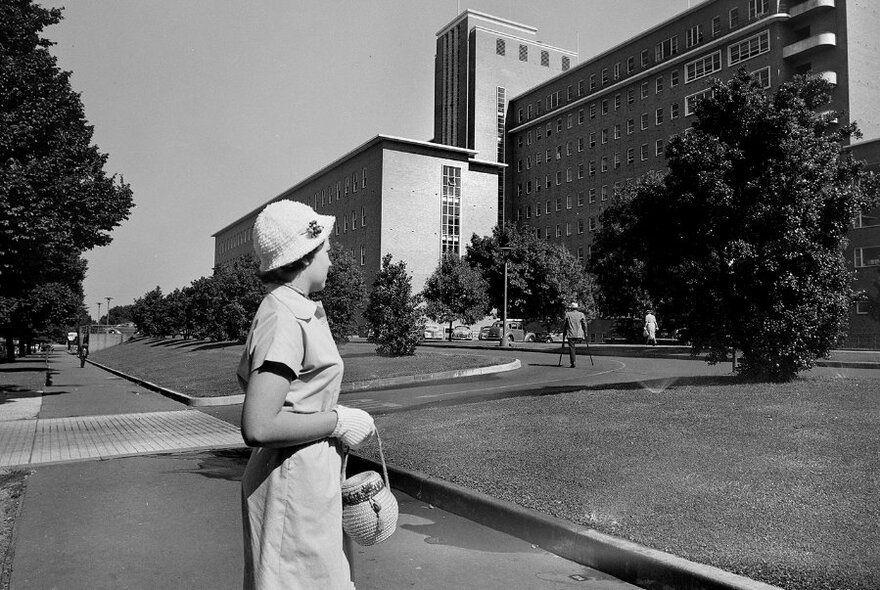 Vintage image of a woman in a uniform and hat crossing a footpath, with lawn, trees and an imposing large building in the background; black and white.