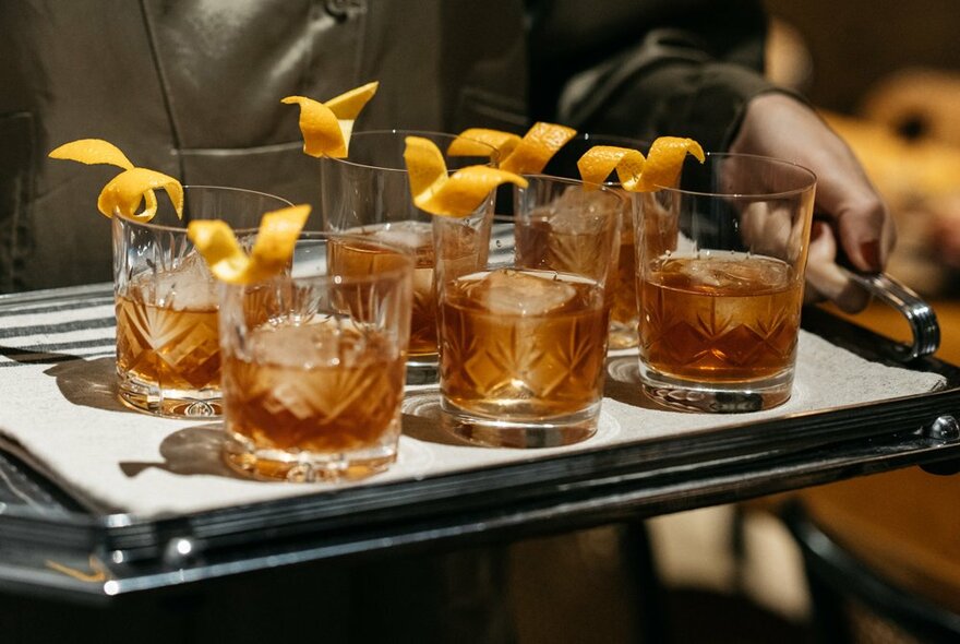 Hands carrying a tray of cocktails served in old fashioned glasses (low tumblers), each with a twist of orange peel garnish.