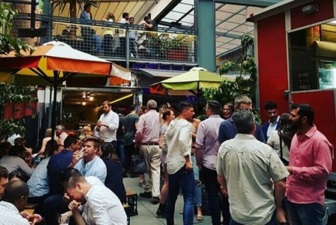 Busy bar area with people standing with drinks and seated at tables under umbrellas.