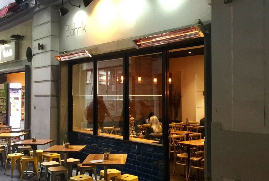 Cafe exterior: tables and stools outside, with ad couple of diners seated inside a cosy interior.
