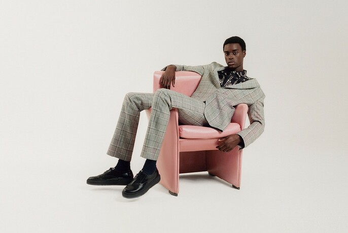 Male model posing on a pink chair, wearing a casual beige suit.