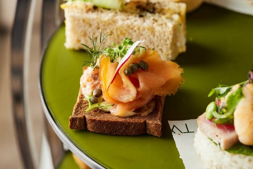 Small piece of bread with salmon and green garnish, finger sandwich to rear.