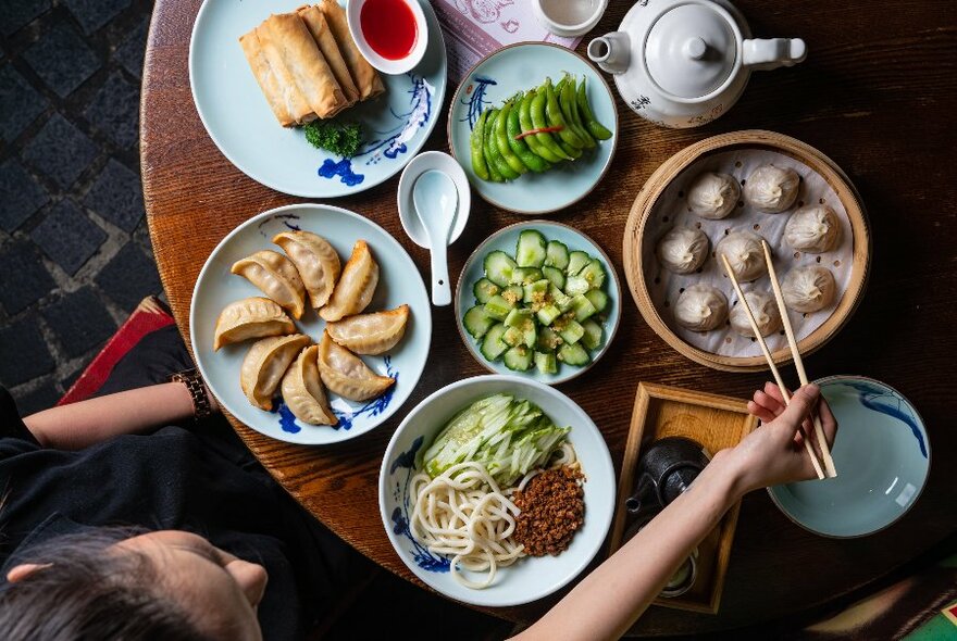 Hands with chopsticks selecting a dumpling from a bamboo steamer, an assortment of food in plates and bowls also on the round table, viewed from above.