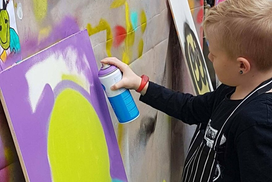Child using a spray can to paint on a canvas in an outdoor area.