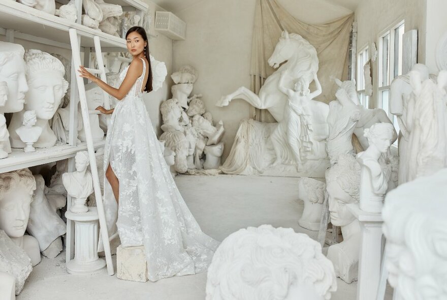 A woman in a long white bridal gown posing on a ladder in a room with classical sculptures.