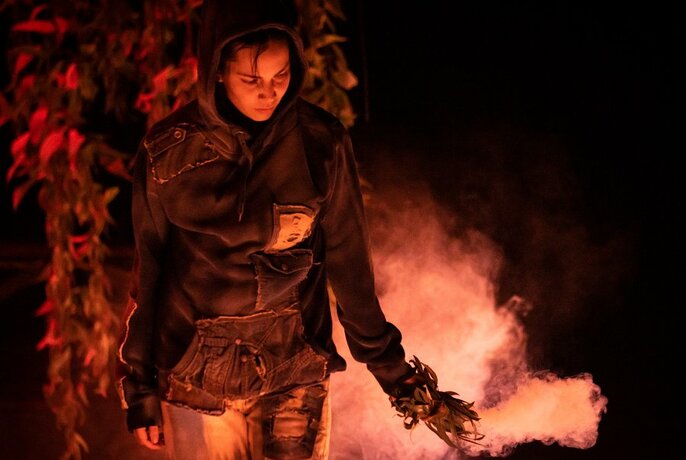 Person wearing dark clothes, looking down, holding smouldering bunch of leaves in one hand and moving them around, smoke visible against the dark background.