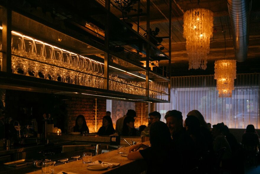 Moody restaurant interior with people seated at a bar with oerhead rows of glasses, lit by stalactite-like a,ber chandeliers.