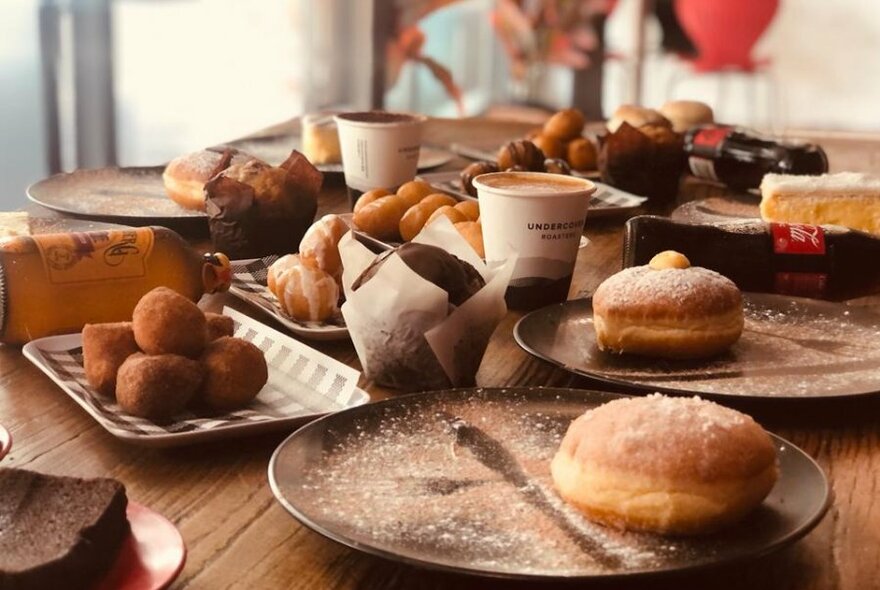 A dining table with plates of pastries and doughnuts, muffins and paper cups of coffee.