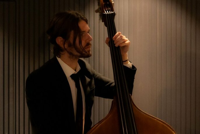 A musician with a long fringe playing the cello, wearing a suit and tie.