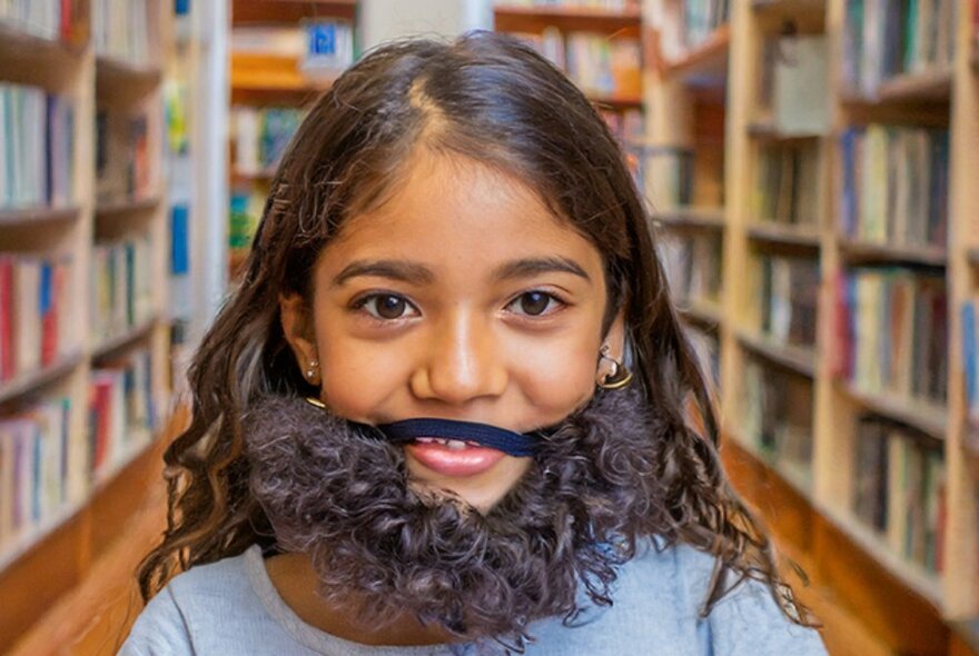 Primary school aged girl standing in a library, between two rows of bookshelves, with long brown hair and a smiling face, wearing a fake beard.