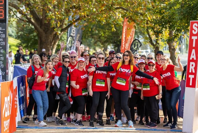 Group of people in matching red t-shirts lined up rand posing at the official start line of a running path, large green leafy trees in background.