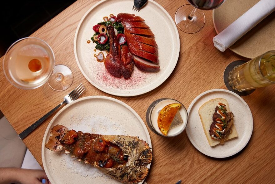 Wooden table with white plates of food including duck, lobster tail and cocktails.