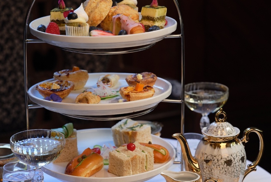 A high tea stand with sandwiches and pastries, a ceramic tea pot and cups and glasses of champagne. 