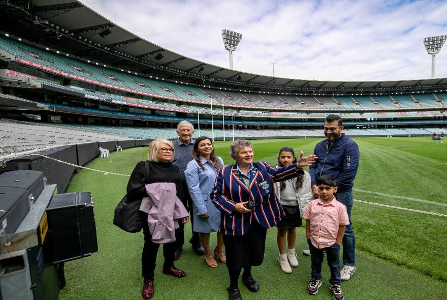 A small group of people standing inside the Melbourne Cricket Ground listening to a woman in a striped blazer as she points out things with her hand. 