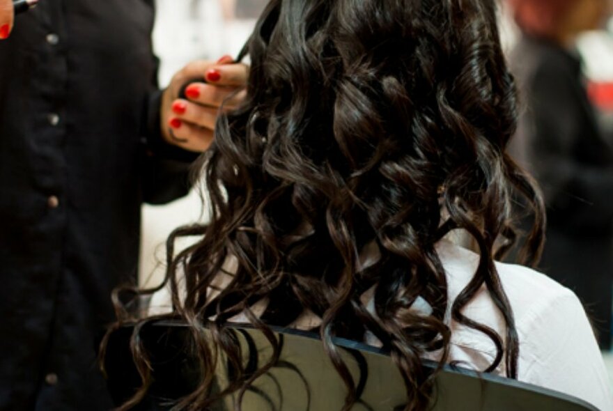 Salon beautician adding long wavy hair extensions to a client seated in a chair.