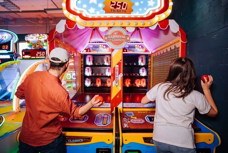 Two people with their backs to the viewer throwing a soft ball at an arcade style 'knock them down' game.