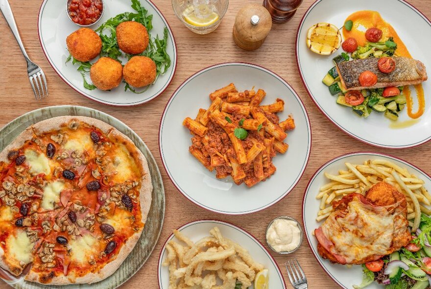 A table with many plates of Italian food including pizza, arancini, pasta, salmon, parmigiana and calamari.