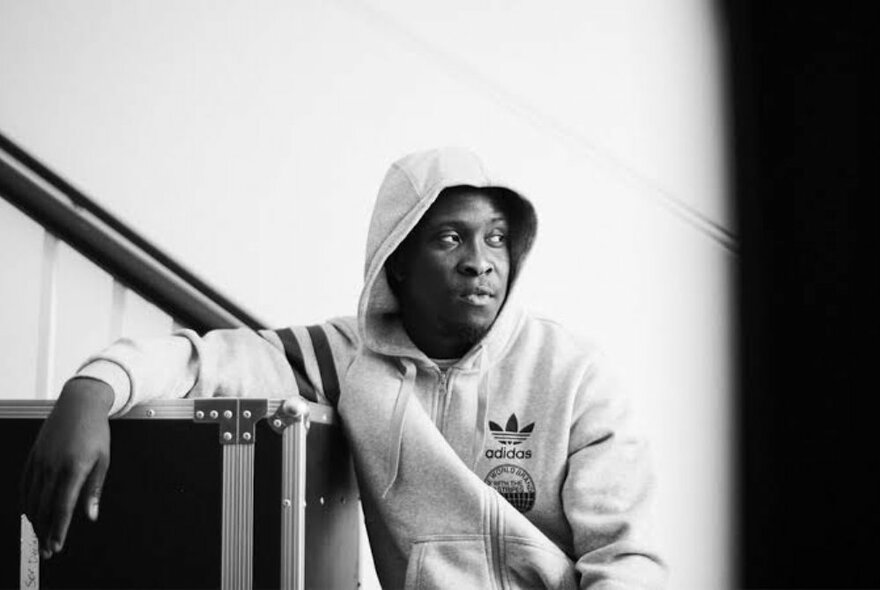 Black and white shot of English musician, Kojo Funds, wearing a light-coloured hooded jumper, leaning against an amplifier.
